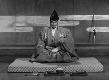 Movie still from “Throne of Blood” (1957), directed by Akira Kurosawa – A man sitting on the ground wearing a hat; Wide shot, High angle