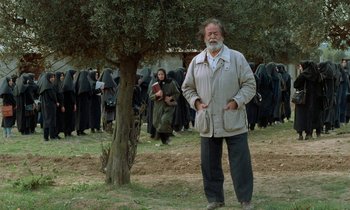 Movie still from “Through the Olive Trees” (1994), directed by Abbas Kiarostami – An old man standing in front of a group of people; Wide shot, Low angle
