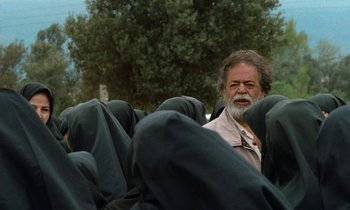Movie still from “Through the Olive Trees” (1994), directed by Abbas Kiarostami – An older man sitting on a chair in front of a tree; Medium shot, Over the shoulder angle