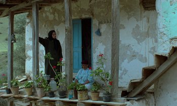 Movie still from “Through the Olive Trees” (1994), directed by Abbas Kiarostami – Two women sitting on a porch with potted plants; Wide shot, Low angle