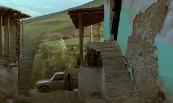Movie still from “Through the Olive Trees” (1994), directed by Abbas Kiarostami – A man standing on the side of a building next to a truck; Extreme Wide shot, Low angle