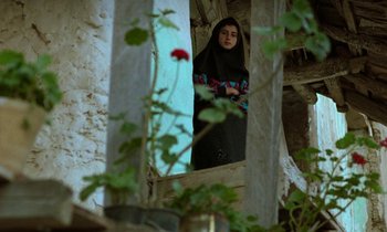 Movie still from “Through the Olive Trees” (1994), directed by Abbas Kiarostami – A woman standing in front of a window with flowers in front of it; Medium shot, Low angle