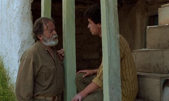 Movie still from “Through the Olive Trees” (1994), directed by Abbas Kiarostami – A man and a man sitting next to each other in front of a brick wall; Medium shot, Over the shoulder angle