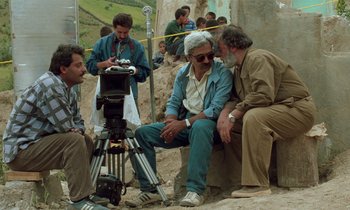 Movie still from “Through the Olive Trees” (1994), directed by Abbas Kiarostami – A group of people sitting around a camera; Wide shot, Over the shoulder angle
