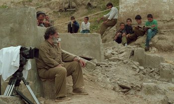 Movie still from “Through the Olive Trees” (1994), directed by Abbas Kiarostami – An old man sitting on a rock with a group of people sitting around him; Wide shot, Over the shoulder angle