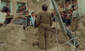 Movie still from “Through the Olive Trees” (1994), directed by Abbas Kiarostami – A group of people sitting on the side of a building; Wide shot, Over the shoulder angle