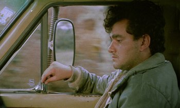 Movie still from “Through the Olive Trees” (1994), directed by Abbas Kiarostami – A man sitting in the driver's seat of a car; Close Up shot, Low angle