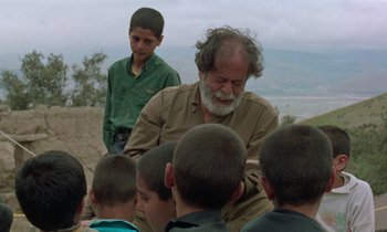 Movie still from “Through the Olive Trees” (1994), directed by Abbas Kiarostami – An older man sitting in front of a group of young boys; Medium shot, Over the shoulder angle