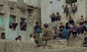 Movie still from “Through the Olive Trees” (1994), directed by Abbas Kiarostami – A group of people sitting and standing around a building; Wide shot, High angle