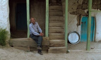 Movie still from “Through the Olive Trees” (1994), directed by Abbas Kiarostami – An older man sitting on a wooden step; Wide shot, Low angle