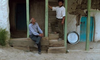 Movie still from “Through the Olive Trees” (1994), directed by Abbas Kiarostami – Two men are sitting on the steps of a house; Wide shot, Low angle