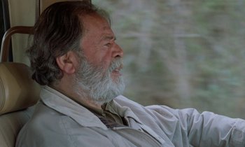 Movie still from “Through the Olive Trees” (1994), directed by Abbas Kiarostami – An older man with a beard is sitting in a chair; Close Up shot, Low angle