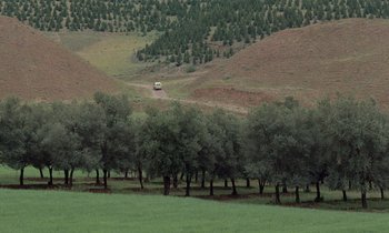 Movie still from “Through the Olive Trees” (1994), directed by Abbas Kiarostami – A bus is driving through a field near some trees; Extreme Wide shot, High angle