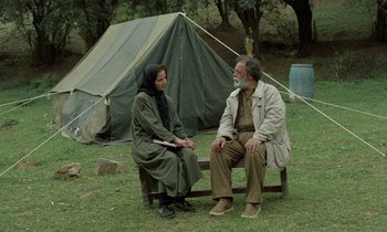 Movie still from “Through the Olive Trees” (1994), directed by Abbas Kiarostami – A man and a woman sitting on a bench in front of a tent; Wide shot, Over the shoulder angle