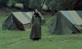 Movie still from “Through the Olive Trees” (1994), directed by Abbas Kiarostami – A woman standing in front of some tents in a field; Wide shot, Low angle