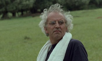 Movie still from “Through the Olive Trees” (1994), directed by Abbas Kiarostami – An older man wearing glasses and a towel around his neck; Close Up shot, Low angle