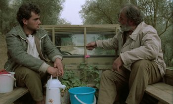 Movie still from “Through the Olive Trees” (1994), directed by Abbas Kiarostami – Two men sitting next to each other near a car; Medium shot, Low angle