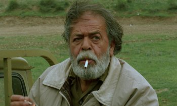 Movie still from “Through the Olive Trees” (1994), directed by Abbas Kiarostami – An older man with a beard and a cigarette in his mouth; Close Up shot, Low angle