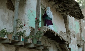 Movie still from “Through the Olive Trees” (1994), directed by Abbas Kiarostami – A woman standing on a balcony looking out the window; Wide shot, Low angle