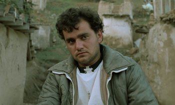 Movie still from “Through the Olive Trees” (1994), directed by Abbas Kiarostami – A person wearing a jacket near a stone wall; Close Up shot, Low angle