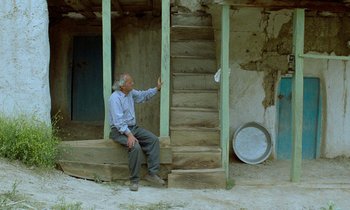 Movie still from “Through the Olive Trees” (1994), directed by Abbas Kiarostami – An older man sitting on a porch next to stairs; Wide shot, Low angle
