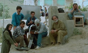Movie still from “Through the Olive Trees” (1994), directed by Abbas Kiarostami – A group of men sitting on the side of a building; Wide shot, High angle