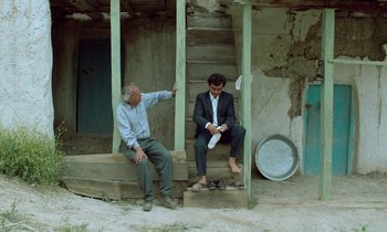 Movie still from “Through the Olive Trees” (1994), directed by Abbas Kiarostami – Two men sitting on steps in front of a building; Wide shot, Low angle