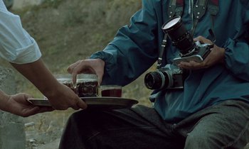 Movie still from “Through the Olive Trees” (1994), directed by Abbas Kiarostami – A man holding a plate with a camera on top of it; Medium shot, Over the shoulder angle