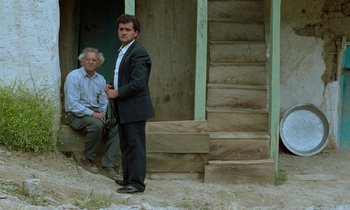 Movie still from “Through the Olive Trees” (1994), directed by Abbas Kiarostami – A man in a suit standing next to another man in a suit; Wide shot, Low angle