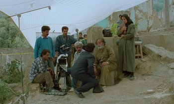 Movie still from “Through the Olive Trees” (1994), directed by Abbas Kiarostami – A group of people standing around a man with a camera; Wide shot, High angle