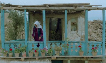 Movie still from “Through the Olive Trees” (1994), directed by Abbas Kiarostami – An old woman standing on a porch of an old house; Wide shot, Low angle