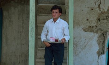 Movie still from “Through the Olive Trees” (1994), directed by Abbas Kiarostami – A man holding a tray of food in front of stairs; Medium shot, Low angle