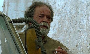 Movie still from “Through the Olive Trees” (1994), directed by Abbas Kiarostami – An older man smoking a cigarette while holding an instrument; Close Up shot, Low angle