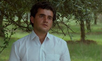 Movie still from “Through the Olive Trees” (1994), directed by Abbas Kiarostami – A man wearing a white shirt standing next to a tree; Close Up shot, Low angle