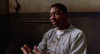 Movie still from “Throw Momma from the Train” (1987), directed by Danny DeVito – A man sitting in front of a brick wall talking; Close Up shot, Over the shoulder angle