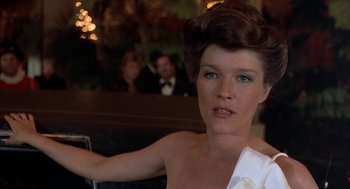 Movie still from “Throw Momma from the Train” (1987), directed by Danny DeVito – A woman in a white dress sitting in front of a crowd; Close Up shot, Low angle