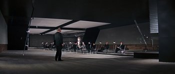 Movie still from “Thunderball” (1965), directed by Terence Young – A group of people sitting in chairs in front of a man in a black suit; Extreme Wide shot, High angle