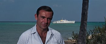 Movie still from “Thunderball” (1965), directed by Terence Young – A man standing in front of the ocean with a boat in the background; Medium shot, Low angle