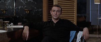 Movie still from “Thunderball” (1965), directed by Terence Young – A man in a black shirt sitting on a couch; Medium shot, Low angle