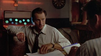 Movie still from “Thunderheart” (1992), directed by Michael Apted – A man sitting at a table with papers in front of him; Medium shot, Over the shoulder angle