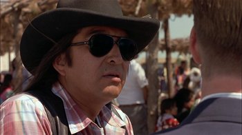 Movie still from “Thunderheart” (1992), directed by Michael Apted – A man wearing a hat and sunglasses in front of a group of people; Close Up shot, Over the shoulder angle
