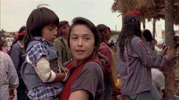 Movie still from “Thunderheart” (1992), directed by Michael Apted – A woman holding a child in her arms while standing in a crowd of people; Close Up shot, Over the shoulder angle