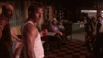 Movie still from “Tin Cup” (1996), directed by Ron Shelton – A man standing in front of another man in a living room; Medium shot, Over the shoulder angle