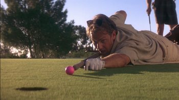 Movie still from “Tin Cup” (1996), directed by Ron Shelton – A man is laying in the grass with a pink ball; Medium shot, Low angle