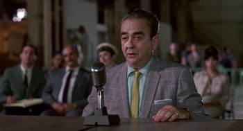 Movie still from “Tin Men” (1987), directed by Barry Levinson – An older man is sitting at a table with a microphone in front of him; Medium shot, Over the shoulder angle