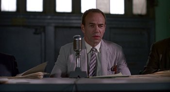 Movie still from “Tin Men” (1987), directed by Barry Levinson – A man sitting at a table in front of a microphone; Medium shot, Low angle