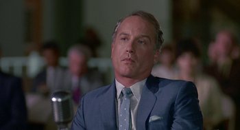 Movie still from “Tin Men” (1987), directed by Barry Levinson – A man in a suit and tie sitting in front of a microphone; Close Up shot, Over the shoulder angle