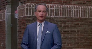 Movie still from “Tin Men” (1987), directed by Barry Levinson – A man in a suit and tie standing in front of a brick wall; Medium shot, Low angle