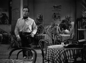 Movie still from “To Have and Have Not” (1944), directed by Howard Hawks – A black and white photo of a man standing in front of a table; Wide shot, Over the shoulder angle