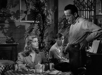 Movie still from “To Have and Have Not” (1944), directed by Howard Hawks – A man and a woman sitting at a table with another man standing next to them; Medium shot, Over the shoulder angle
